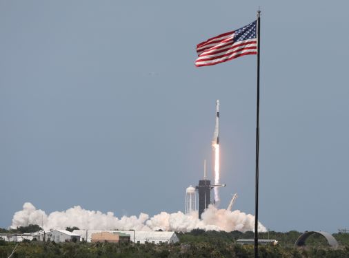 A SpaceX Falcon 9 rocket lifts off, framed by an American flag atop a flagpole.