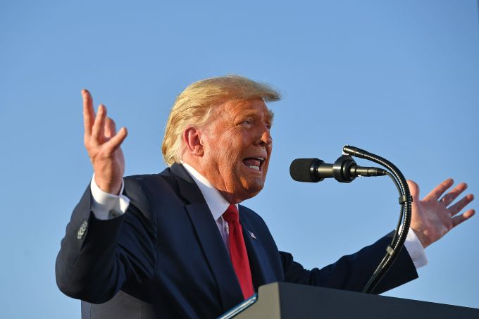 Donald Trump speaking into a microphone against a backdrop of the sky. He is gesticulating with his hands.