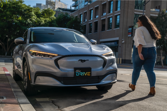 Woman walks up to silver Mustang Mach-E. License plate says "Ford Drive"