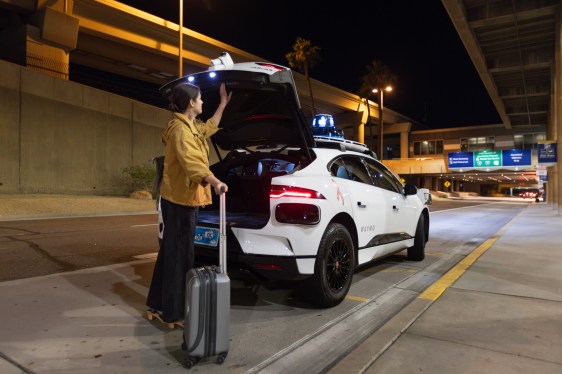 A man stands behind a Waymo robotaxi with the trunk open. One hand goes to close the trunk, the other rests on his luggage.
