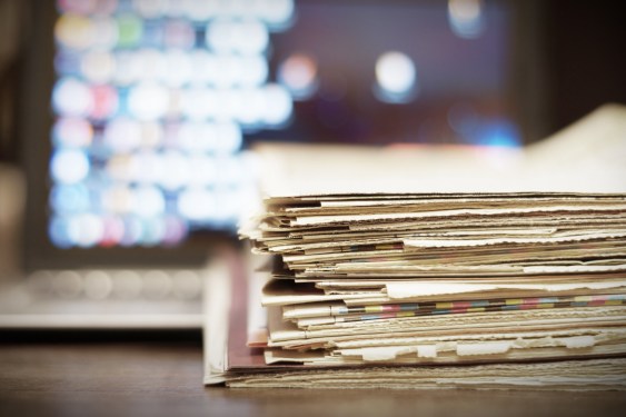 a stack of newspapers and documents in front of a blurry display of a laptop
