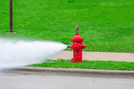 Red fire hydrant shooting water in front of sidewalk and bright green lawn.