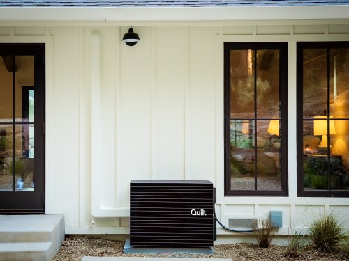A Quilt heat pump outdoor unit sits outside a white house.